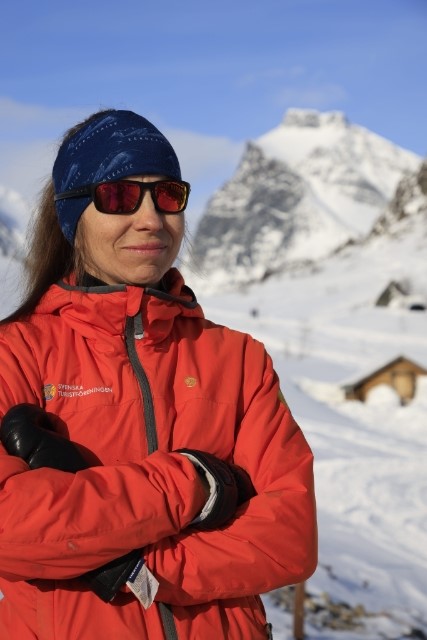 Marit Sarri i röd teckjacka med snöklädda fjäll i bakgrunden.